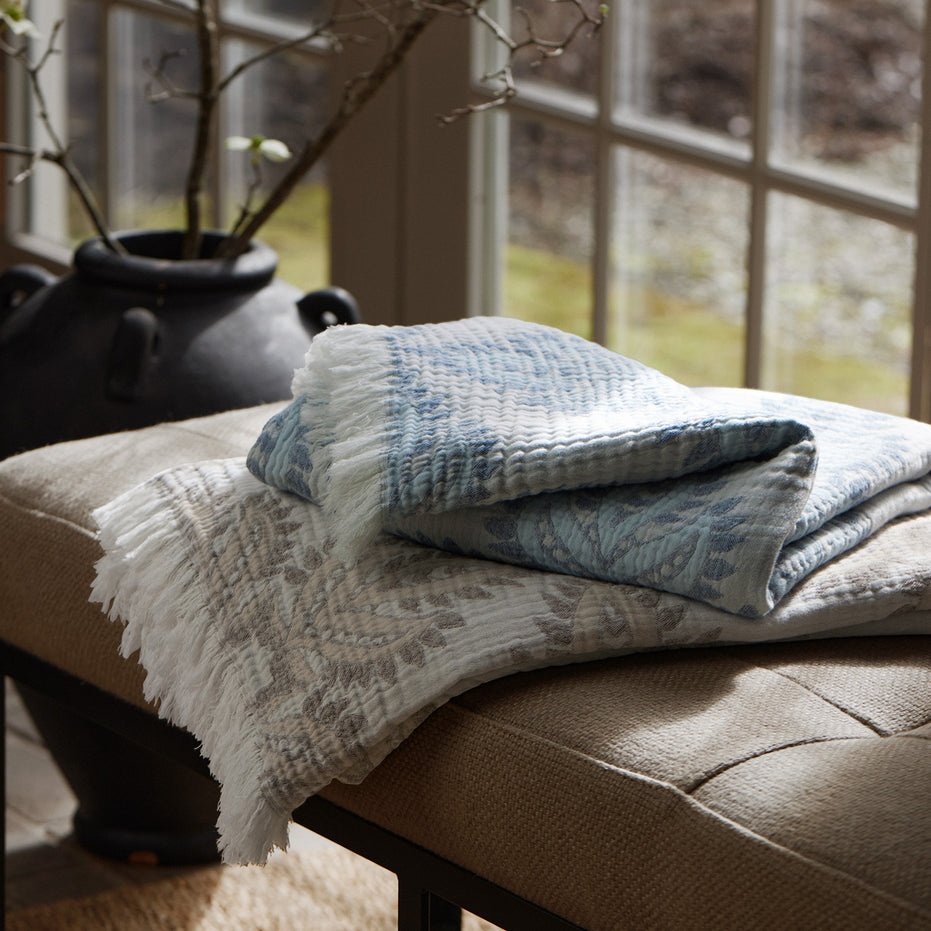 Two folded Dasati Sand throws by John Robshaw, featuring stonewashed cotton with intricate woven designs and fringed edging, are placed on a cushioned bench in front of a window, with a black vase in the background.