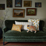 A dark green couch with four patterned pillows and a stylish John Robshaw Farah Marigold Decorative Pillow sits elegantly. One of the cushions features hand-stitched edging, adding a touch of craftsmanship. The wall behind the couch is adorned with framed paintings, while a tall branch with white flowers stands to the left. - 31259422261294