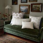 A dark green velvet couch with various patterned cushions, including the Nivi Euro embroidered pillow of the John Robshaw brand featuring a hidden zipper closure, sits in front of a wall adorned with framed paintings. A side table with a floral arrangement is next to the couch. - 31263122456622