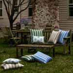 Backyard scene featuring weathered wooden furniture, including a table, chairs, and benches, adorned with John Robshaw's Lucy Moss Outdoor Kidney Pillows made of hand block printed cotton linen. A potted plant with purple flowers sits on the table. - 31261349150766