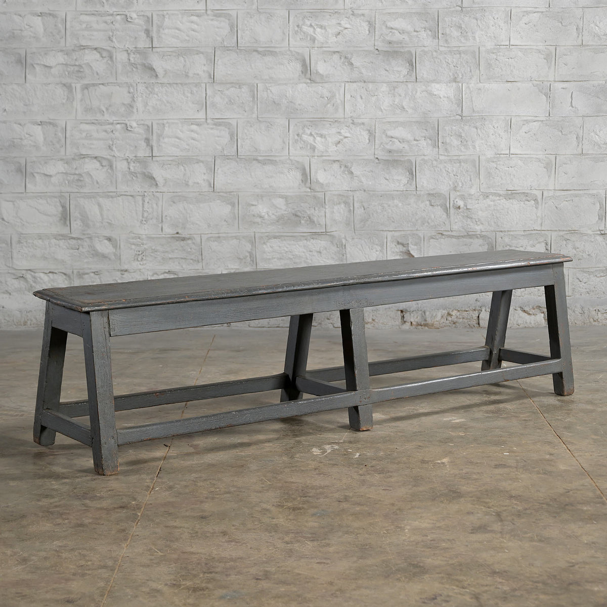 A long, dark gray Madura Teak Bench by John Robshaw, featuring angled legs, sits on a concrete floor against a textured white brick wall, evoking the timeless character of classic school benches.