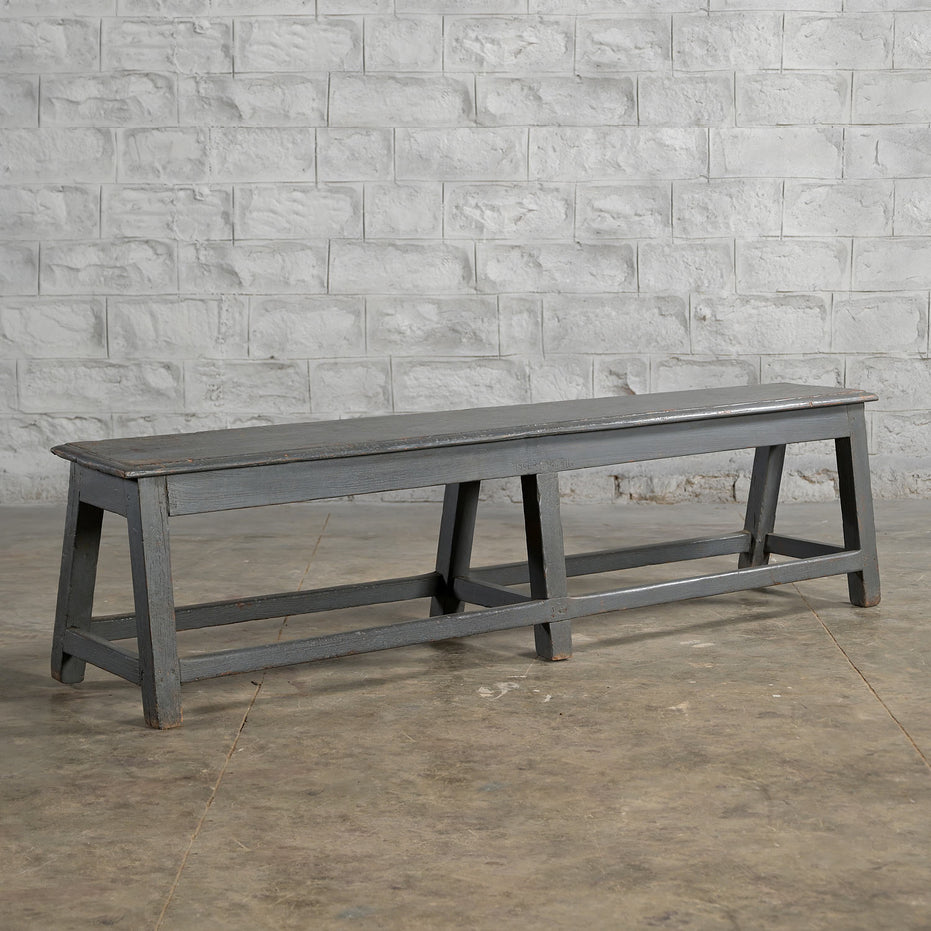 A long, dark gray Madura Teak Bench by John Robshaw, featuring angled legs, sits on a concrete floor against a textured white brick wall, evoking the timeless character of classic school benches.