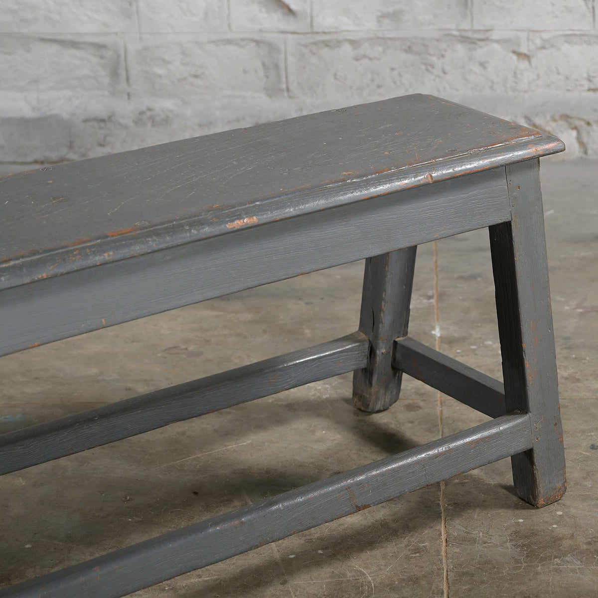 A close-up of the Madura Teak Bench by John Robshaw, featuring its weathered gray finish, set on a concrete floor with a white brick wall in the background.
