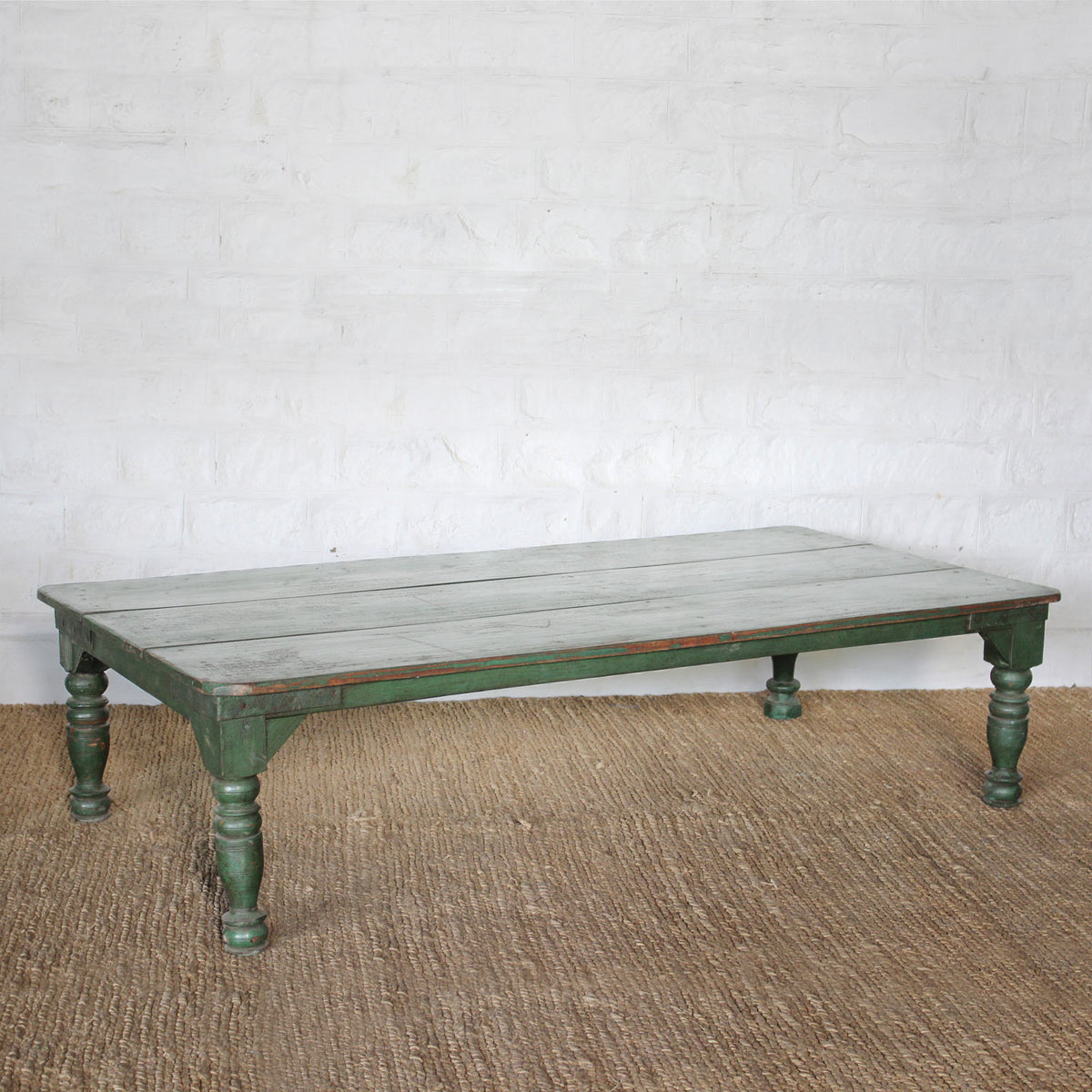 Aangan Teak Coffee Table by John Robshaw, featuring a low rectangular design with weathered green legs and a whitewashed plank-style top, displayed on a woven rug against a white brick wall.