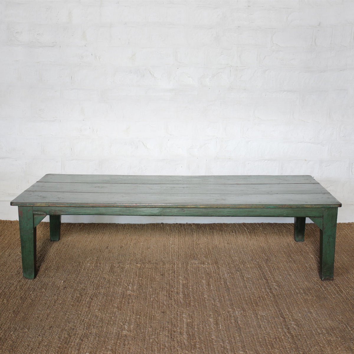 Aangan Teak Coffee Table by John Robshaw sits on a woven rug against a white textured brick wall, featuring a simple silhouette with a weathered green base and a gray plank-style top.