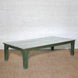 The Aangan Teak Coffee Table by John Robshaw features a low, rectangular design with a weathered green frame and a light-colored, plank-style top, shown placed on a textured brown rug against a white brick wall.