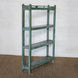 The Paarthiv Wooden Shelf by John Robshaw, featuring a weathered green finish with a subtle teal patina and three shelves, stands on a brown woven rug against a white textured wall, offering a unique touch of vintage charm to the space.