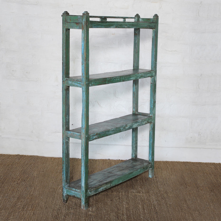 The Paarthiv Wooden Shelf by John Robshaw, featuring a weathered green finish with a subtle teal patina and three shelves, stands on a brown woven rug against a white textured wall, offering a unique touch of vintage charm to the space.