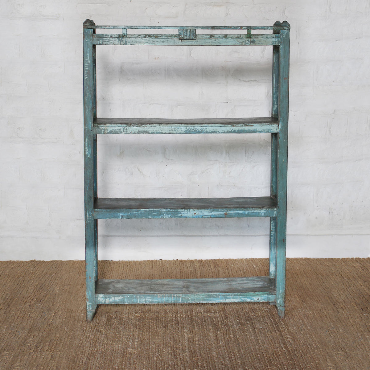 The Paarthiv Wooden Shelf by John Robshaw, with its distressed blue finish and subtle teal patina, stands against a white brick wall on a brown woven rug, exuding the timeless charm of vintage furniture.