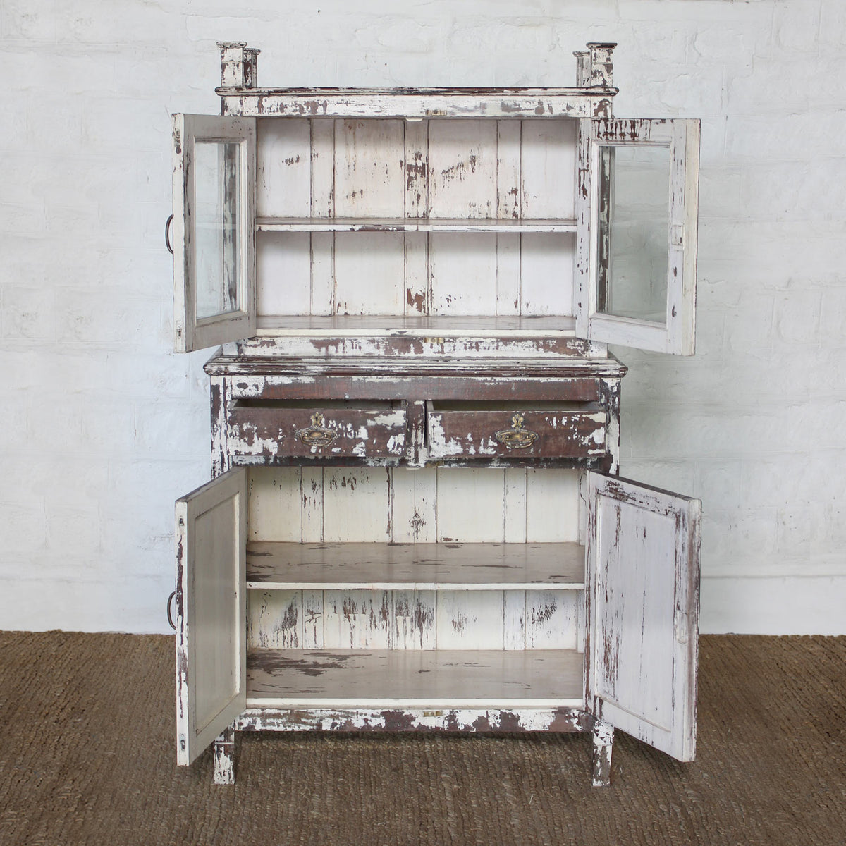 Introducing the Juhi Teak Cabinet by John Robshaw—a character-rich piece crafted in distressed white wood. It features two glass-paneled upper doors, two drawers, and two lower doors, all opening to reveal generous storage and shelving within.
