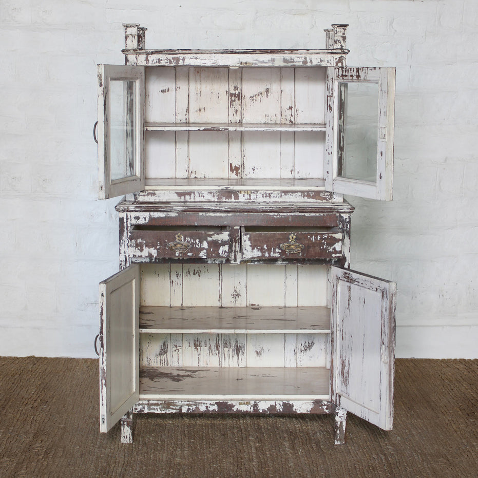 Introducing the Juhi Teak Cabinet by John Robshaw—a character-rich piece crafted in distressed white wood. It features two glass-paneled upper doors, two drawers, and two lower doors, all opening to reveal generous storage and shelving within.