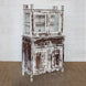 The Juhi Teak Cabinet by John Robshaw is a character-rich piece featuring a distressed wooden finish, glass-paneled doors on top, two drawers, and two solid doors below for ample storage. Standing gracefully on a woven rug against a white brick wall, it brings both charm and functionality to any space.