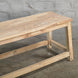 The Umed Teak Bench by John Robshaw, featuring a simple handcrafted design and natural finish, stands on a concrete floor against a textured gray brick wall.