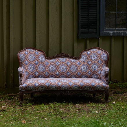 The Maansi Chestnut Sofa by John Robshaw, featuring an elegant camelback silhouette and vintage pattern, sits on the grass in front of a green wooden building with a window and shutter.