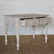 The Jharna Teak Desk by John Robshaw, featuring two open drawers, turned legs, and a distressed finish, sits on a woven rug against a white brick wall, bringing vintage furniture charm to the space.
