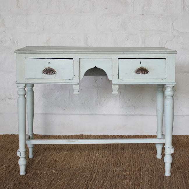 The Pravah Teak Desk by John Robshaw features a light blue vintage finish, two drawers with ornate brass cup pulls, and stands on four turned legs. It is shown placed on a woven rug against a white brick wall.