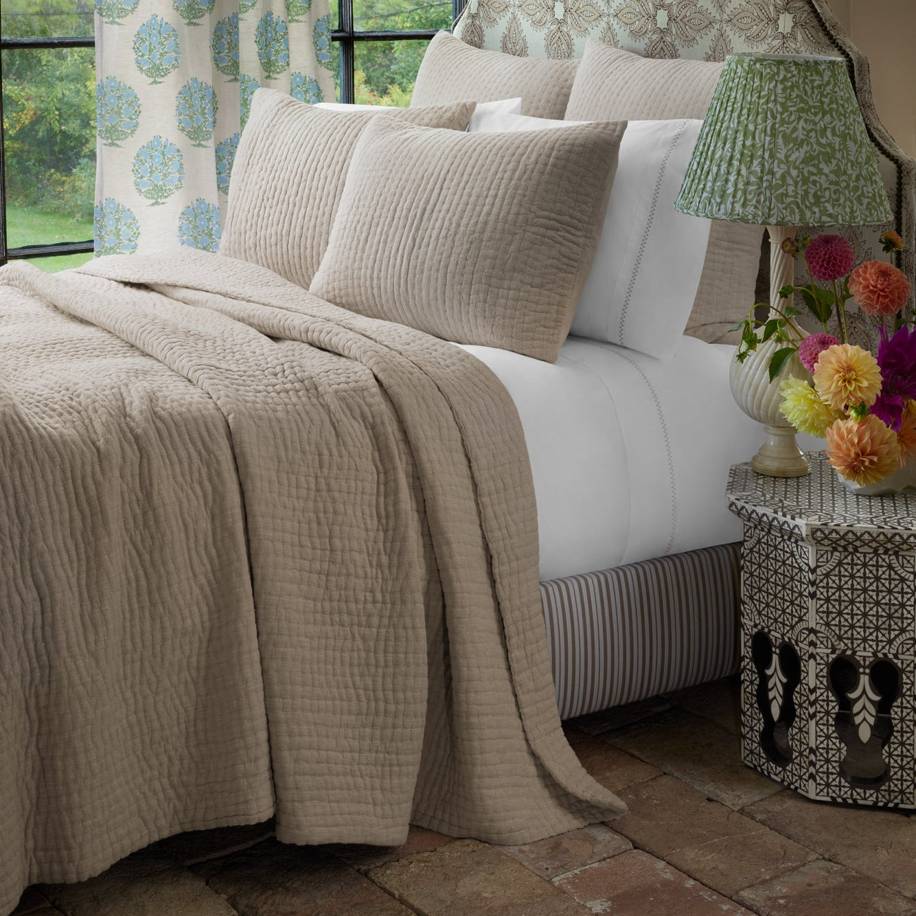 A bed dressed with a beige John Robshaw Vivada Sand Woven Sham, a matching quilted throw, crisp white cotton sheets, a green patterned lamp, and floral curtains beside a window.