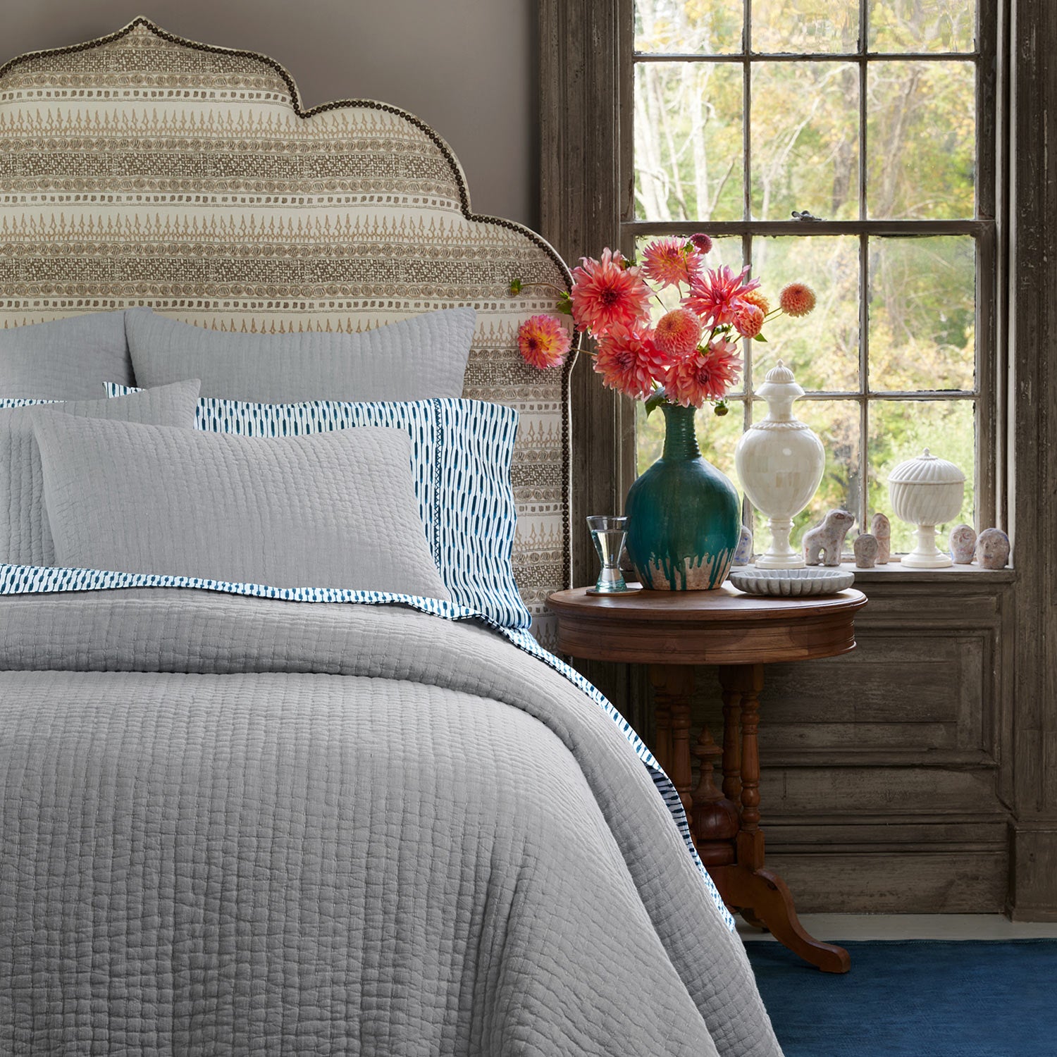 A John Robshaw Vivada Light Gray Woven Quilt with blue and white cotton chambray coverlets, and a window.