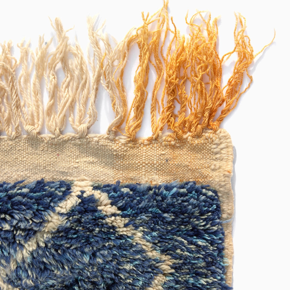 Corner of a Blue Village Wool Rug in textured blue, cream, and yellow wool with long, fringed edges.