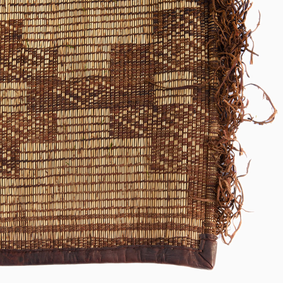 John Robshaw Tazarine Tuareg Mat, a handwoven palm reed mat featuring a geometric brown pattern, leather-bound edges, and a fringed side.