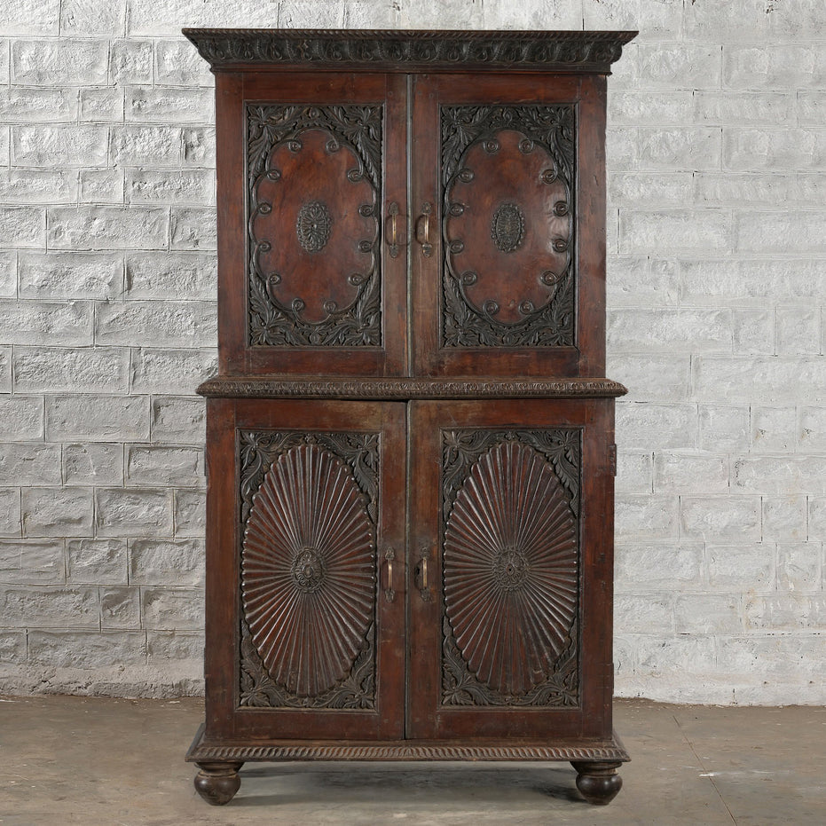 The Bhuv Teak 2 Part Cabinet by John Robshaw features ornate carved doors with circular motifs and decorative trim, displayed against a white textured brick wall on a concrete floor. Showcasing exquisite carved detailing throughout, this piece exemplifies vintage furniture craftsmanship.
