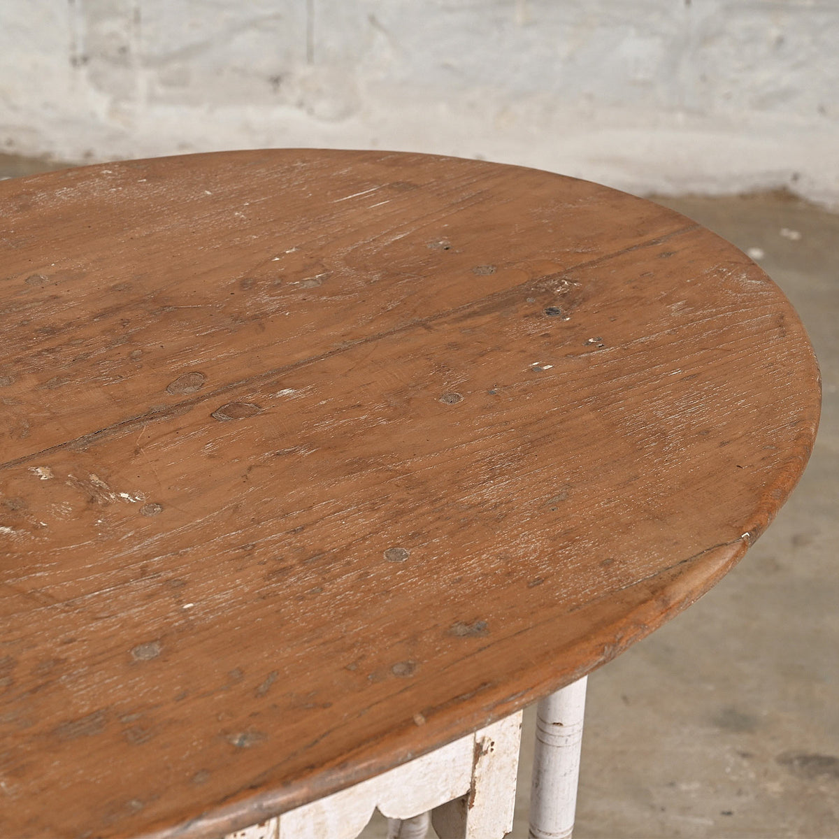 The John Robshaw Teak Table 10 features a round, handcrafted teak design with a weathered brown top and visible wood grain. Set against a concrete floor and light brick wall, this table is perfect for those seeking vintage furniture charm.