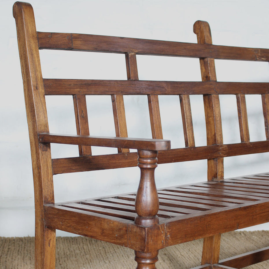 The Shahad Teak Bench by John Robshaw features a vintage design with a slatted seat and turned legs, highlighted by vertical and horizontal slats on the backrest and armrests. It’s shown here set against a white wall atop a woven floor mat.