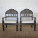 The Himavat Teak Seating Set by John Robshaw features two wooden armchairs with hand-carved backrests and detailed joinery, upholstered in light-colored fabric, placed side by side on a woven rug against a white brick wall.