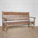 The Mayank Teak Bench by John Robshaw, featuring a slatted back and seat, straight armrests, turned legs, and subtle carved cutout details, is set against a white brick wall on a woven rug.
