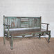 The Pavan Teak Bench by John Robshaw showcases rustic character with its weathered, distressed green finish, slatted backrest, and gracefully curved front legs—perfectly placed on a woven rug against a white brick wall.