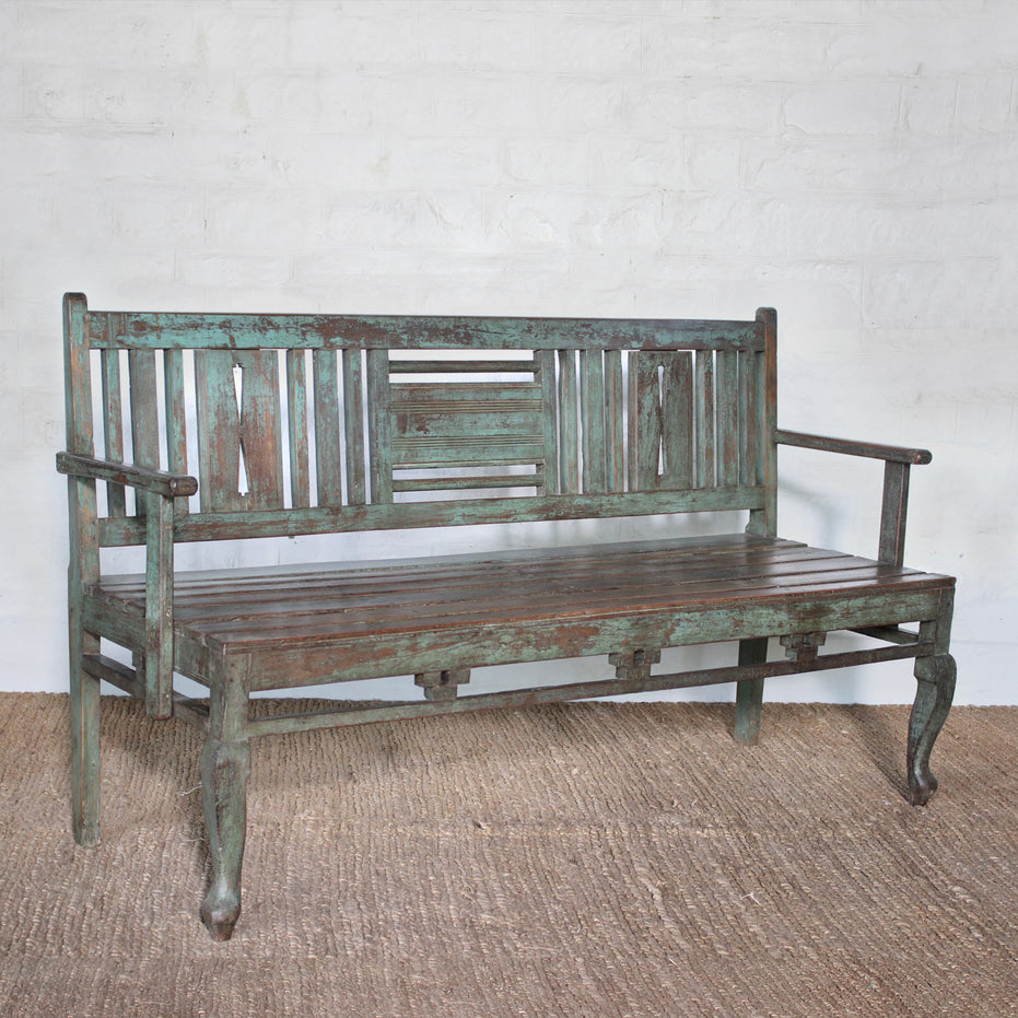 The Pavan Teak Bench by John Robshaw showcases rustic character with its weathered, distressed green finish, slatted backrest, and gracefully curved front legs—perfectly placed on a woven rug against a white brick wall.