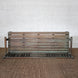 The Pavan Teak Bench by John Robshaw, featuring a weathered finish and rustic character, is set against a white brick wall and positioned on a brown woven rug.