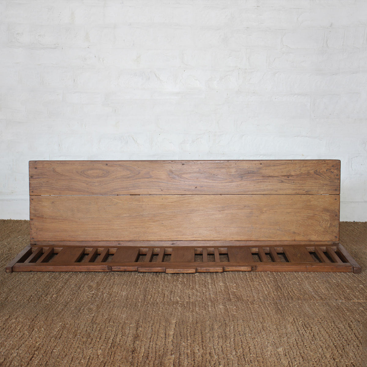 The Sarang Teak Bench by John Robshaw, featuring handcrafted slatted teak seating and a solid backrest, adds the timeless charm of vintage furniture to any space when placed atop a textured brown carpet against a white brick wall.