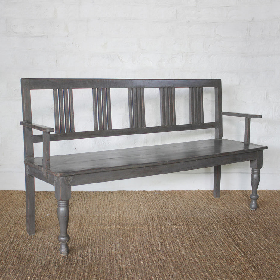 The Varun Teak Bench by John Robshaw, featuring a dark wooden finish with vertical slats on the backrest, armrests, and turned front legs, sits atop a woven rug against a white brick wall, exemplifying the timeless charm of handcrafted vintage furniture.