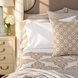 A neatly made bed showcases the Anketi Natural Organic Sheet Set by John Robshaw—plush, 300 thread count organic cotton sheets in beige and white patterns—paired with coordinating pillows and accompanied by a cream nightstand holding a lamp and a vase of white flowers.