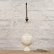 The Shiva Bone Inlay Lamp by John Robshaw features a tall, textured cream-colored ceramic base without a lampshade, evoking the look of a handcrafted piece. It is displayed on a wooden surface in front of a white brick wall, with a clear cord coiled at the base.