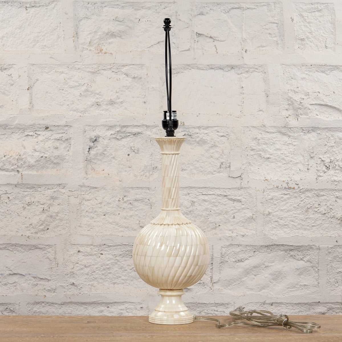 The Shiva Bone Inlay Lamp by John Robshaw features a tall, textured cream-colored ceramic base without a lampshade, evoking the look of a handcrafted piece. It is displayed on a wooden surface in front of a white brick wall, with a clear cord coiled at the base.