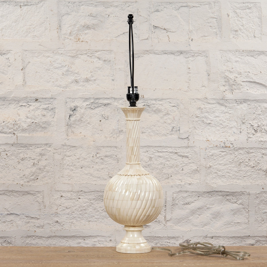 The Shiva Bone Inlay Lamp by John Robshaw features a tall, textured cream-colored ceramic base without a lampshade, evoking the look of a handcrafted piece. It is displayed on a wooden surface in front of a white brick wall, with a clear cord coiled at the base.