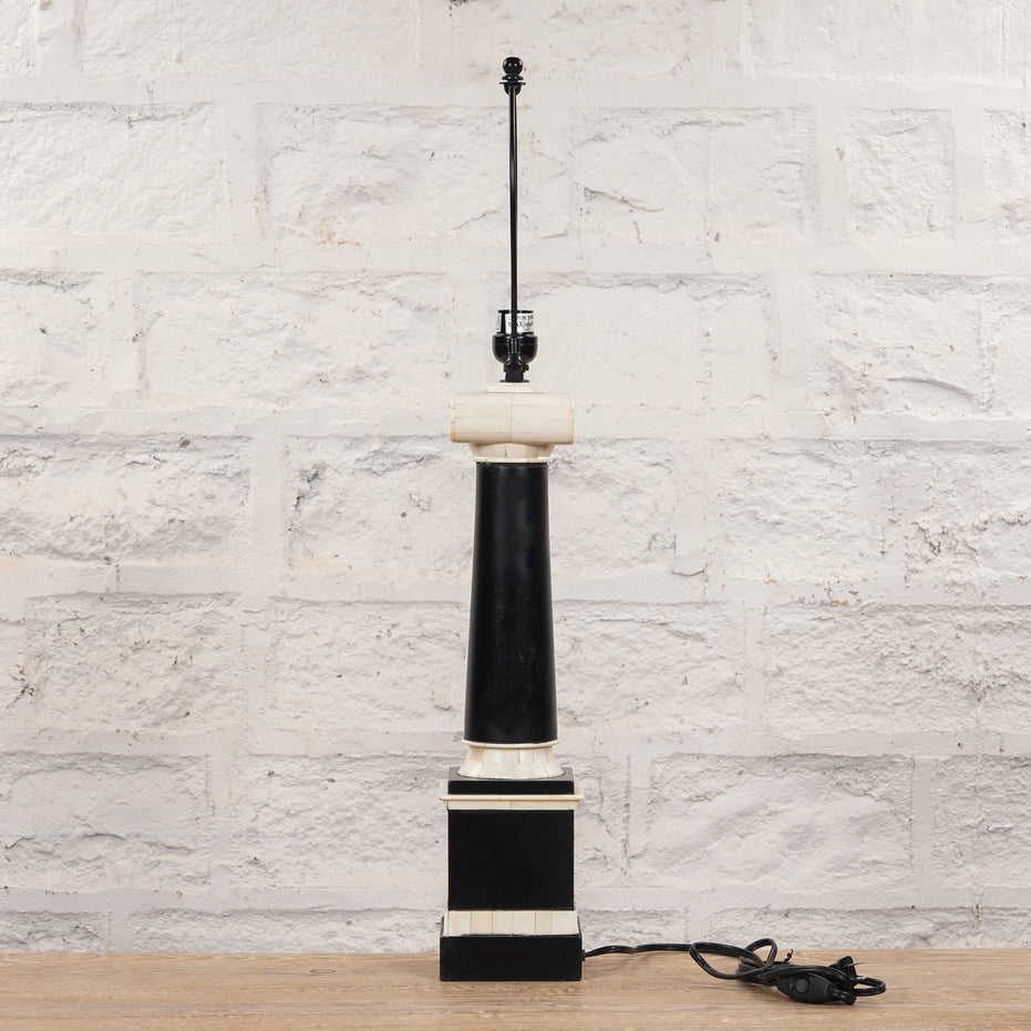 The Roshni Bone Inlay Lamp by John Robshaw, featuring a tall black and white colonial Indian design with a square base, is displayed unplugged on a wooden surface against a textured white brick wall.