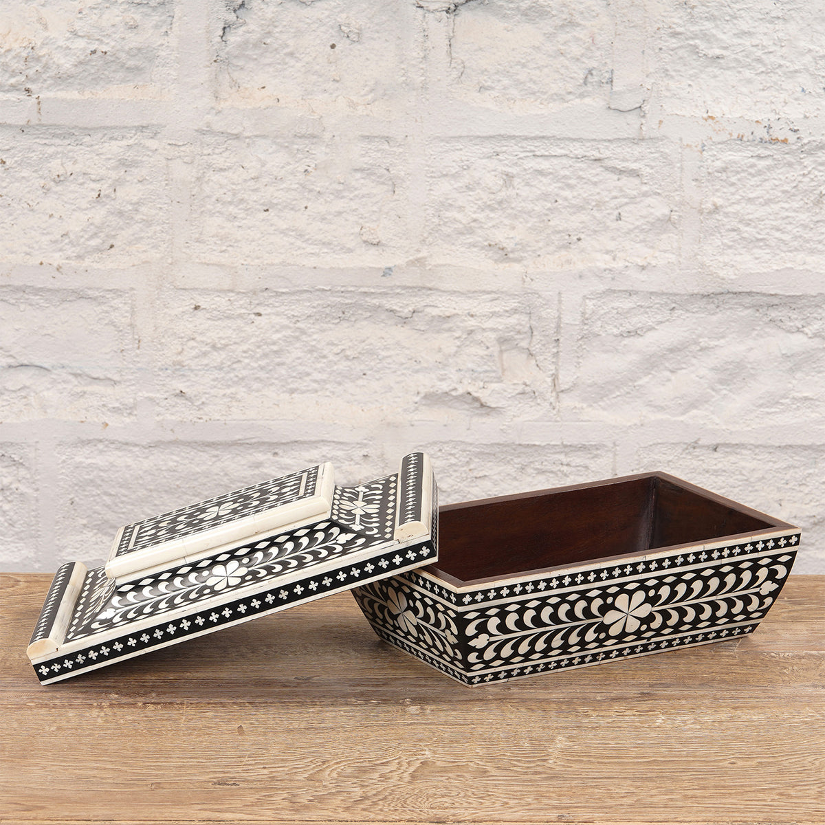 The Rajata Bone Inlay Box by John Robshaw features intricate white floral patterns on a black rectangular surface, displayed with its lid partially open and set atop a wooden surface against a white brick wall.