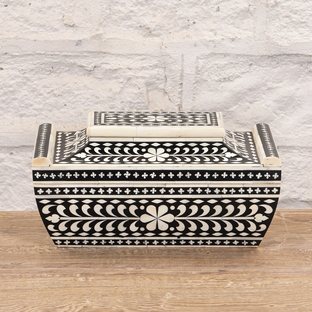 The Rajata Bone Inlay Box by John Robshaw is a rectangular wooden box featuring black and white floral and geometric bone inlay patterns, crafted from Indian teak and displayed on a wooden surface against a light brick wall background.