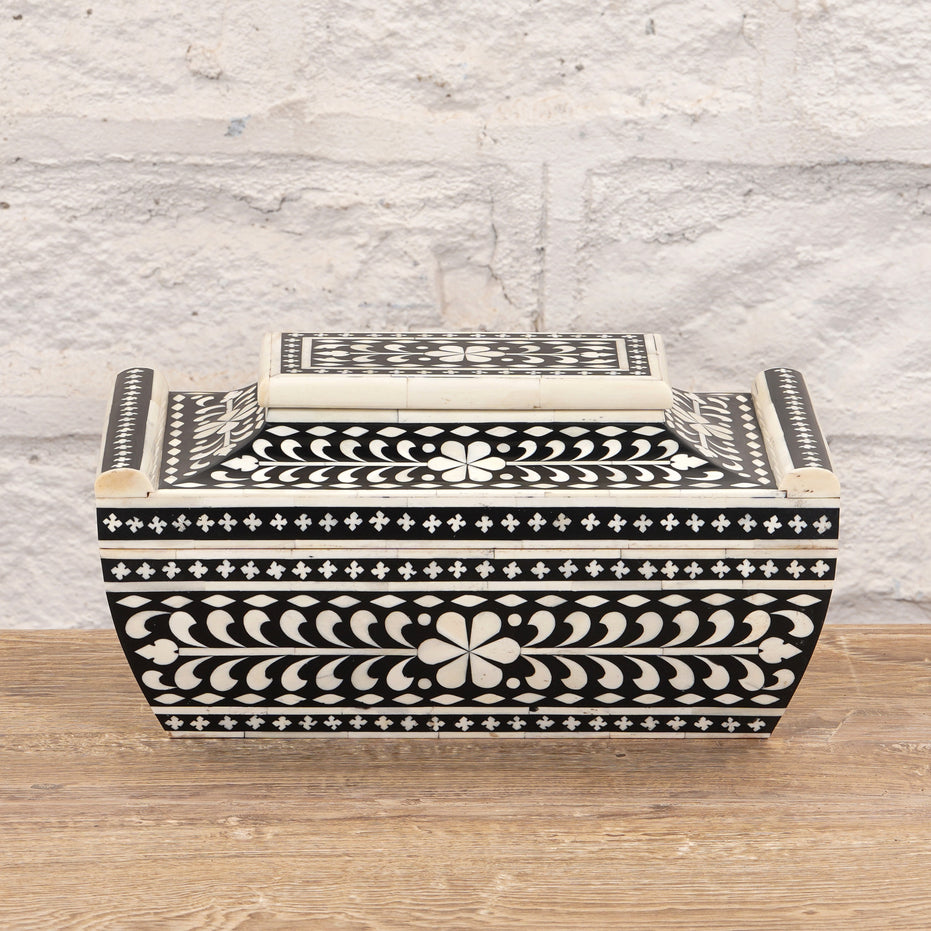 The Rajata Bone Inlay Box by John Robshaw is a rectangular wooden box featuring black and white floral and geometric bone inlay patterns, crafted from Indian teak and displayed on a wooden surface against a light brick wall background.