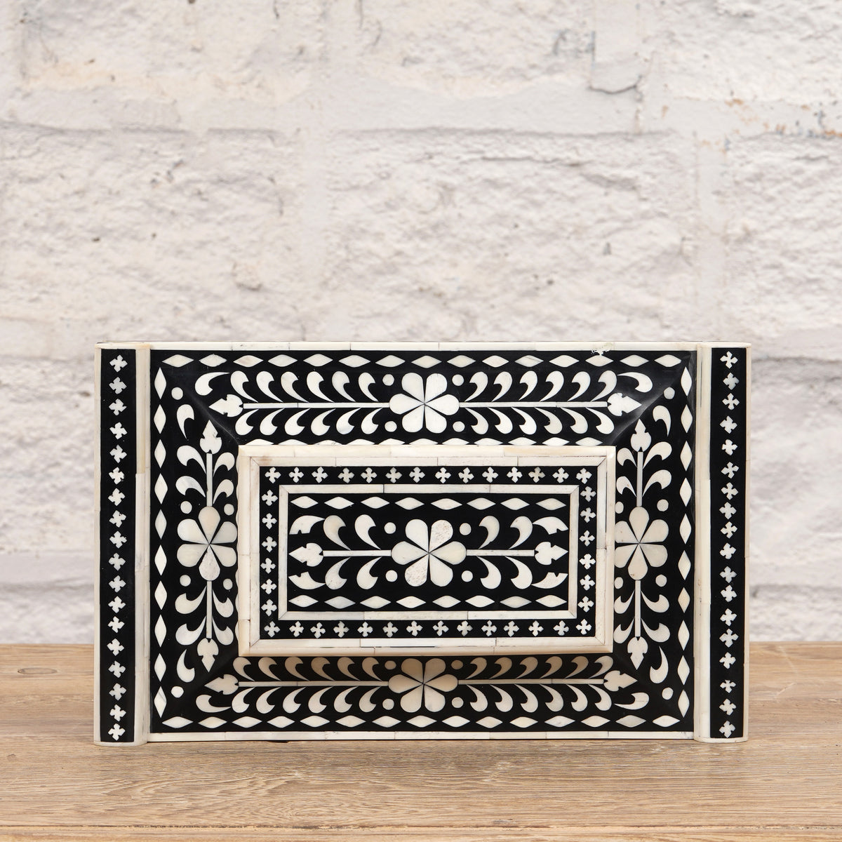 The Rajata Bone Inlay Box by John Robshaw, featuring a rectangular shape and intricate white floral and geometric patterns on a black base, is displayed atop an Indian teak wooden surface with a light brick wall in the background.