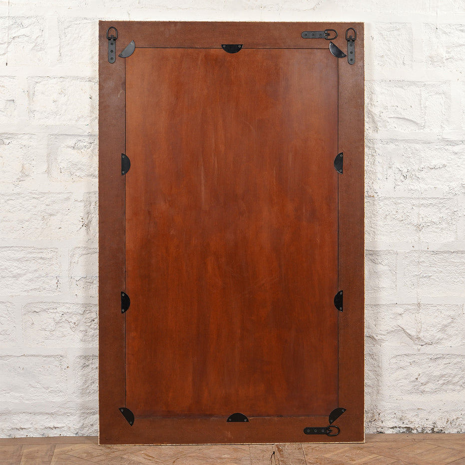 A large rectangular wooden panel with metal brackets and hinges on the edges, leaning against a white brick wall, stands next to the striking Arohi Bone Inlay Mirror by John Robshaw, which features a bold black and white diamond pattern.