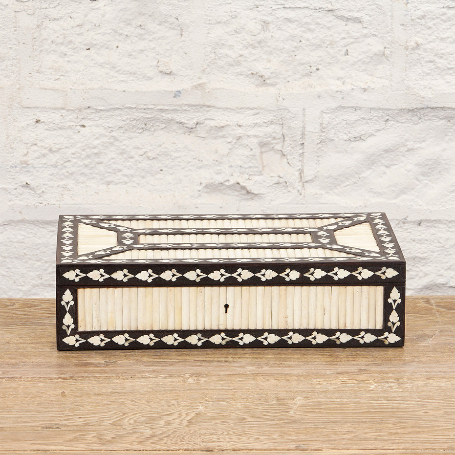 The Charita Bone Inlay Box by John Robshaw, featuring an intricate black and white striped pattern, is placed on a wooden surface against a white brick wall.