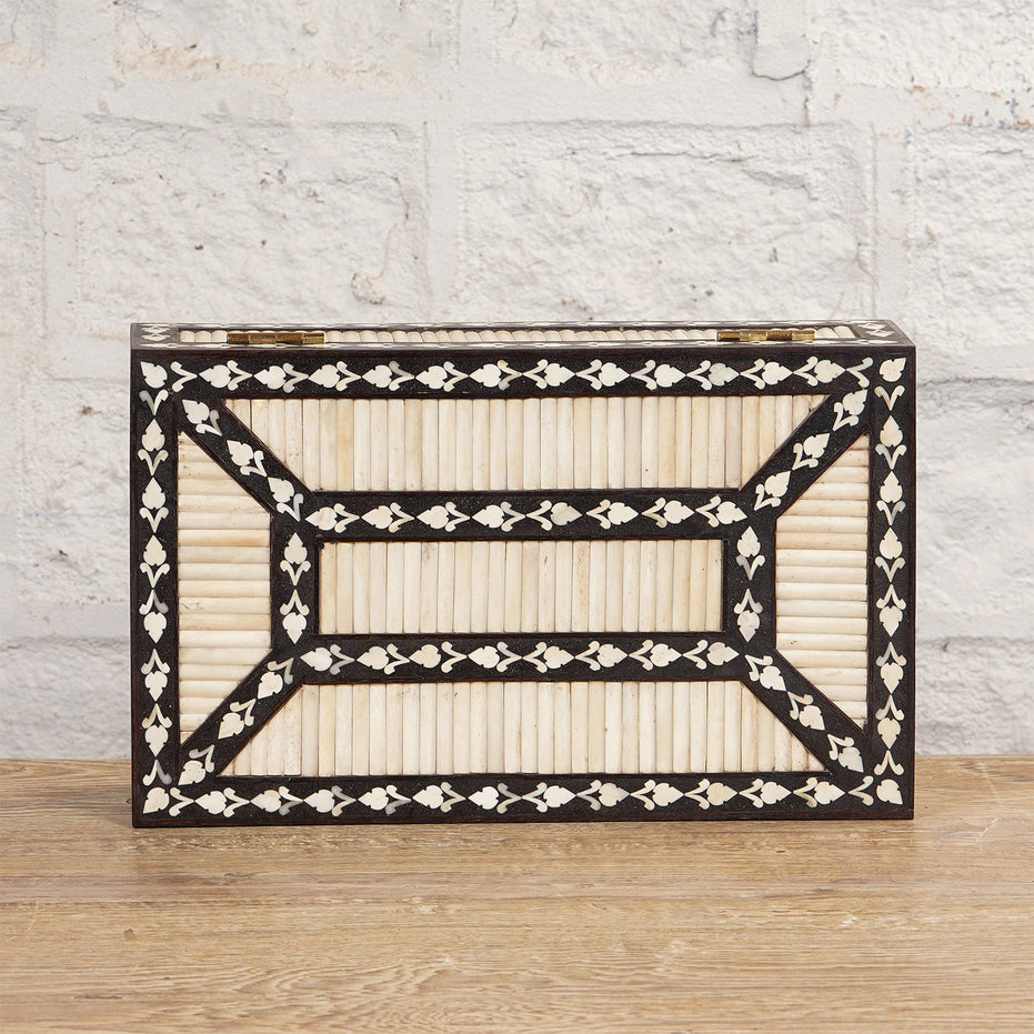 The Charita Bone Inlay Box by John Robshaw, featuring a geometric black and white striped pattern, sits on a teak wooden surface against a white brick wall.