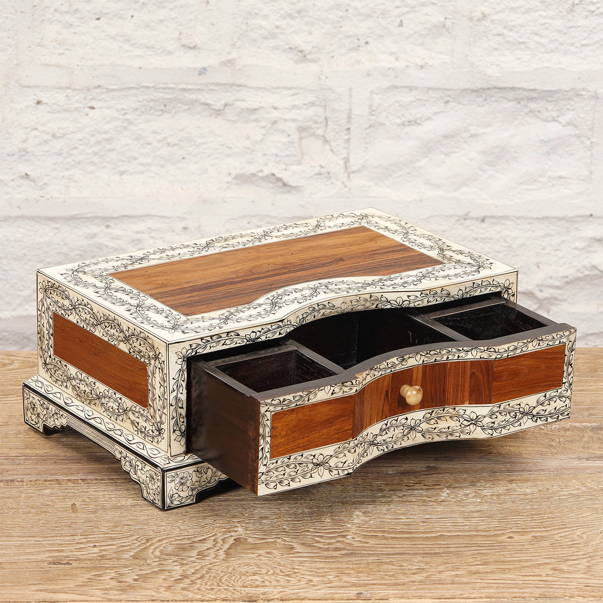 The Harini Bone Inlay Box by John Robshaw is a rectangular wooden jewelry box featuring intricate white floral inlay, three compartments, and an open drawer. It sits elegantly on a wooden surface against a white brick wall.
