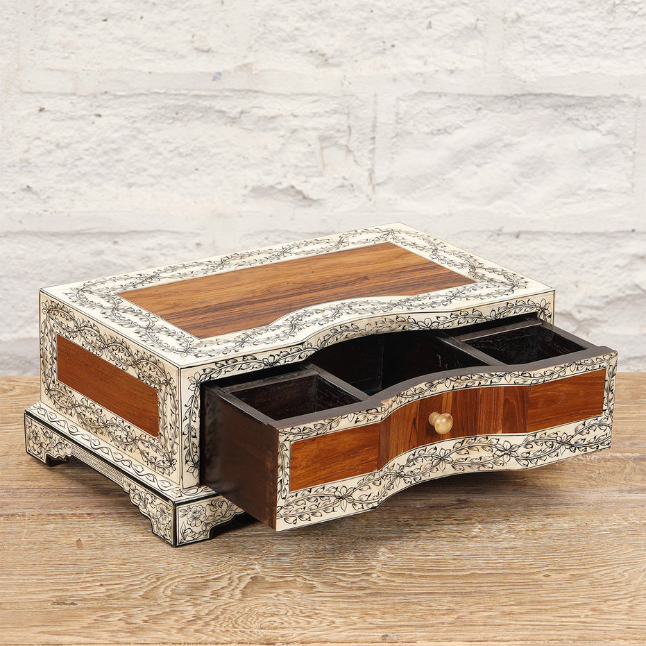 The Harini Bone Inlay Box by John Robshaw is a rectangular wooden jewelry box featuring intricate white floral inlay, three compartments, and an open drawer. It sits elegantly on a wooden surface against a white brick wall.