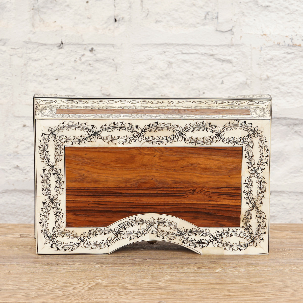 The Harini Bone Inlay Box by John Robshaw is a rectangular decorative wooden piece, handcrafted with intricate vine patterns and a light border, shown here set against a white brick wall and wooden surface.