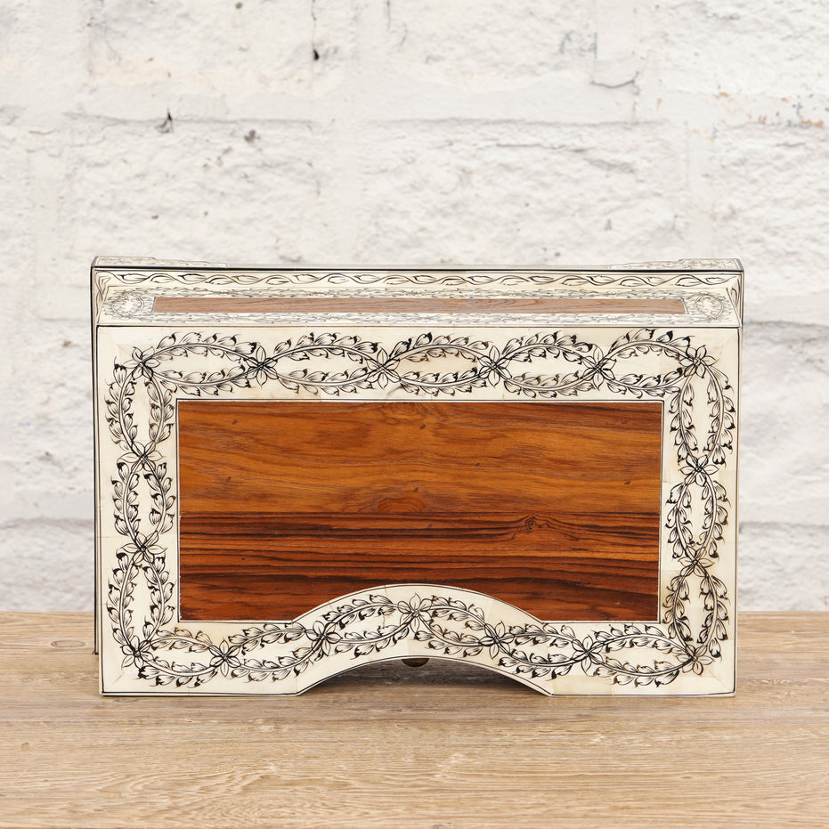 The Harini Bone Inlay Box by John Robshaw is a rectangular decorative wooden piece, handcrafted with intricate vine patterns and a light border, shown here set against a white brick wall and wooden surface.